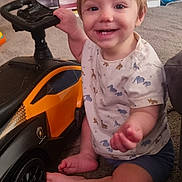 Orlando is registered to the contest to win money with this photo: toddler, child, toy_car, car, smiling, indoor, carpet, happy, shorts, shirt, animal_print, baby, footwear, furniture, sitting, hand, face, head, wheel, playtime