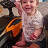 toddler, child, toy_car, car, smiling, indoor, carpet, happy, shorts, shirt, animal_print, baby, footwear, furniture, sitting, hand, face, head, wheel, playtime