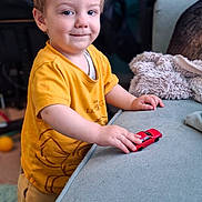 Orlando is registered to the contest to win money with this photo: child, toddler, smiling, portrait, indoor, yellow_shirt, toy_car, red_car, playing, sofa, couch, blanket, home, living_room, seated, standing, short_hair, happy, face, furniture
