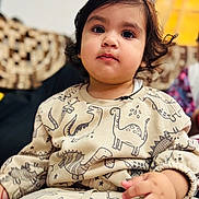 Kevika is registered to the contest to win money with this photo: toddler, child, face, portrait, dinosaur_print, clothing, cute, indoors, sitting, hand, hair, expression, young_child, person, soft_lighting, cozy, patterned_clothes, curly_hair, young, casual