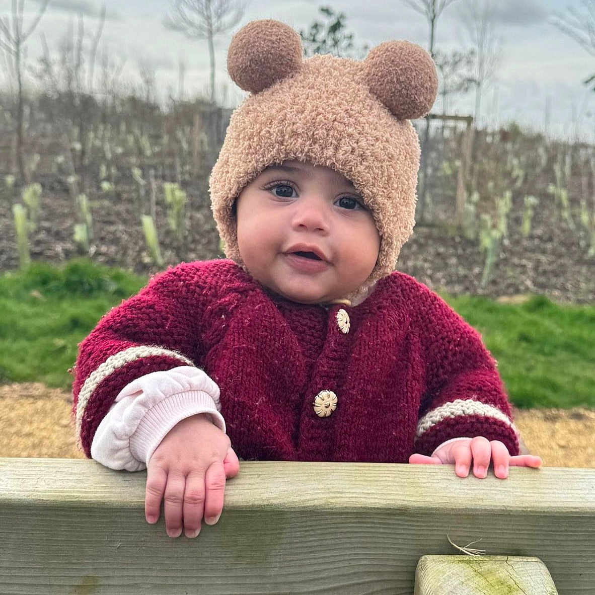Noore is registered to the contest to win money with this photo: baby, bear_ears, bench, button, cheeks, child, cold_weather_clothing, cute, face, grass, hands, knit_hat, outdoor, portrait, red_sweater, sky, smile, toddler, trees, wooden_fence
