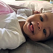 Alana joined the competition — help win amazing prizes! baby, smile, tongue, white_clothing, blanket, bed, pillow, infant, face, cheek, lying_down, cute, expression, skin, head, happy, indoors, soft_light, closeup, person