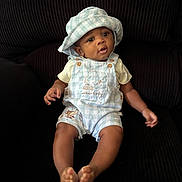 Antonio is registered to the contest to win money with this photo: baby, child, infant, hat, overalls, blue, white, checkered, sitting, couch, dark_background, curious, cute, indoors, clothing, portrait, feet, hands, face, person