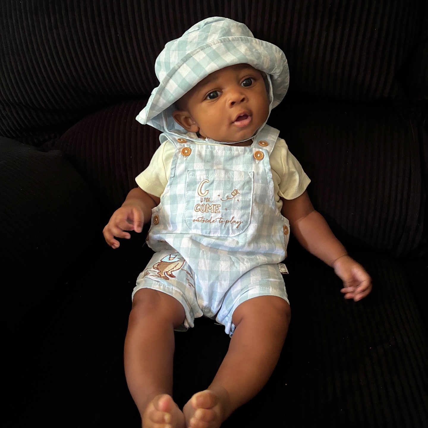 Antonio is registered to the contest to win money with this photo: baby, child, infant, hat, overalls, blue, white, checkered, sitting, couch, dark_background, curious, cute, indoors, clothing, portrait, feet, hands, face, person