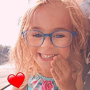 Ashley a rejoint le concours — aidez-le/la à gagner de superbes lots ! child, smiling, glasses, blonde_hair, curly_hair, floral_dress, hand_on_face, window, sunlight, red_heart_emoji, happy, portrait, indoors, person, cute, young_child, casual_clothing, natural_light, bright, closeup
