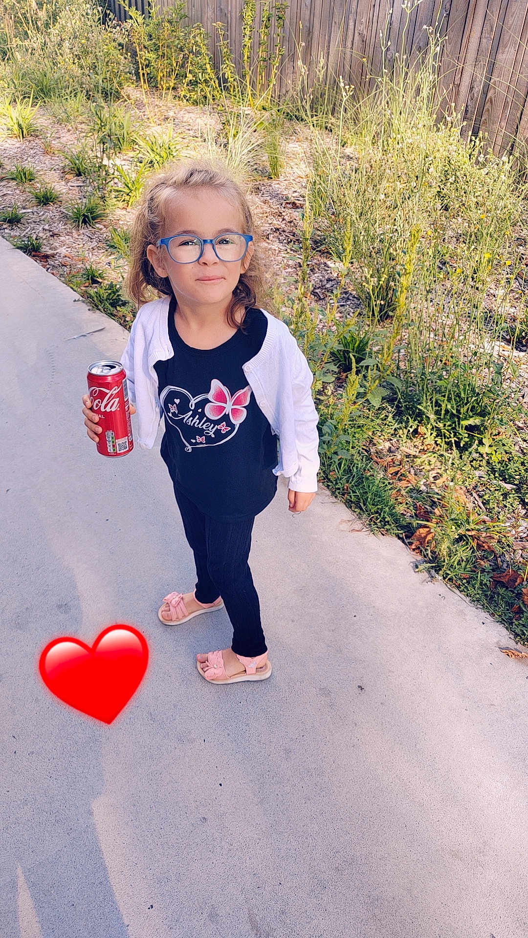 Ashley participe au concours pour gagner de l'argent avec cette photo : child, girl, glasses, blue_glasses, white_cardigan, black_tshirt, butterfly_design, pink_sandals, sidewalk, can_of_soda, outdoor, plants, fence, greenery, sunlight, shadow, heart_emoji, casual_clothing, standing, smiling