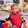Kenzo participe au concours pour gagner de l'argent avec cette photo : toddler, child, outdoor, vest, plaid_shirt, holding_cloth, pavement, trees, chairs, tables, adult_hand, glasses, red, yellow, blue, person, sky, building, sidewalk, casual_clothing