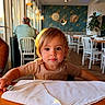 child, toddler, portrait, restaurant, cafe, table, napkin, spoon, seated, indoors, smiling, face, eyes, hair, shirt, warm_lighting, sunlight, chairs, dining_area, plant