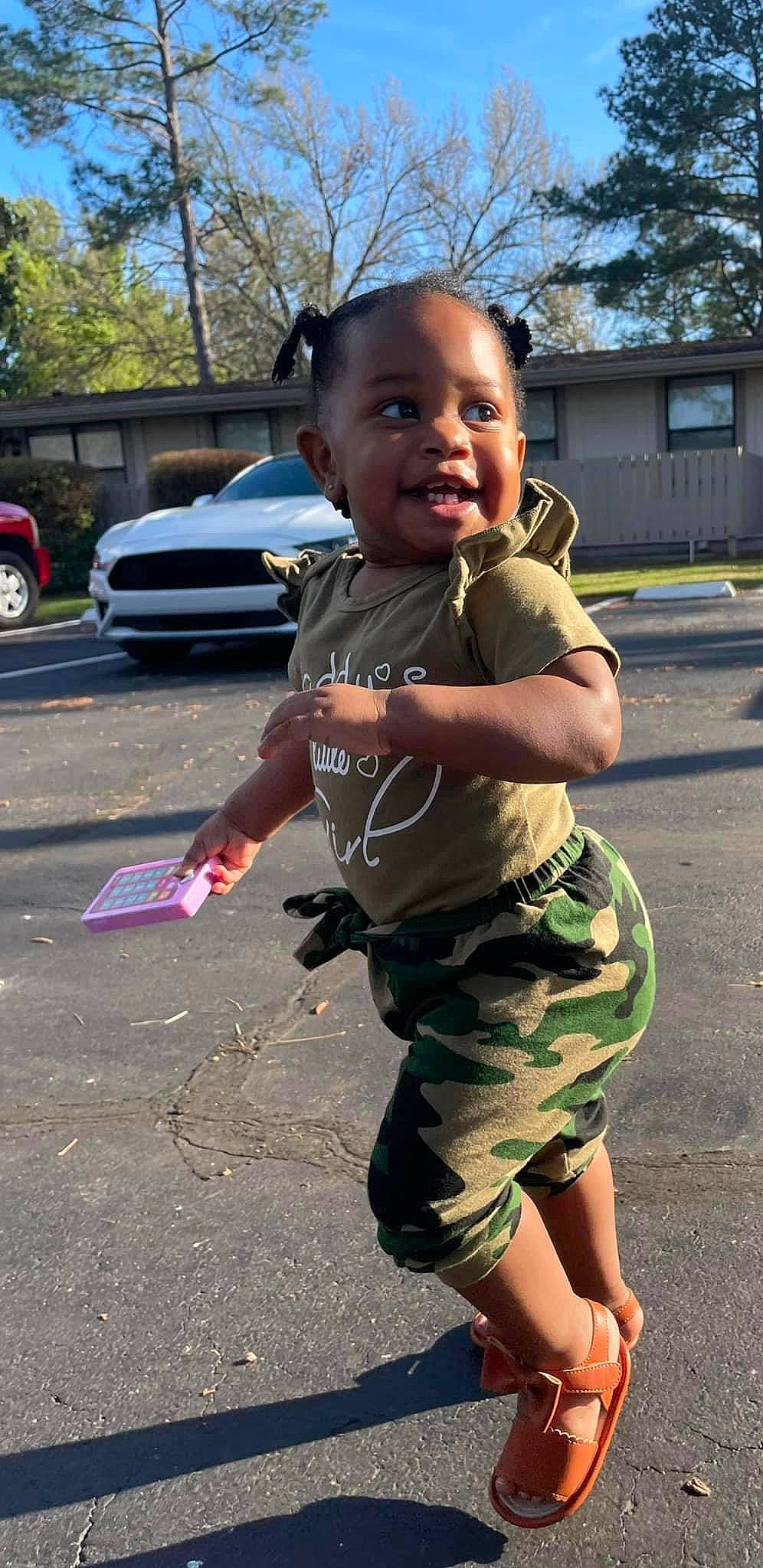 Kylia is registered to the contest to win money with this photo: arm, asphalt, camouflage, car, child, finger, fun, gesture, hand, happy, joint, leg, military_camouflage, person, sleeve, t_shirt, thumb, tire, toddler, tree