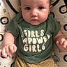 baby, baby_girl, beige_pants, blue_eyes, child, closeup, clothing, cute, expression, face, green_shirt, hands, indoor, infant, lion_pattern, patterned_cushion, portrait, sitting, text_on_clothing, young_child