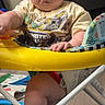 baby, infant, child, portrait, serious_expression, sitting, activity_jumper, yellow, toy, indoor, play_area, graphic_shirt, bare_legs, face, eyes, hands, furniture, home, photograph, colorful