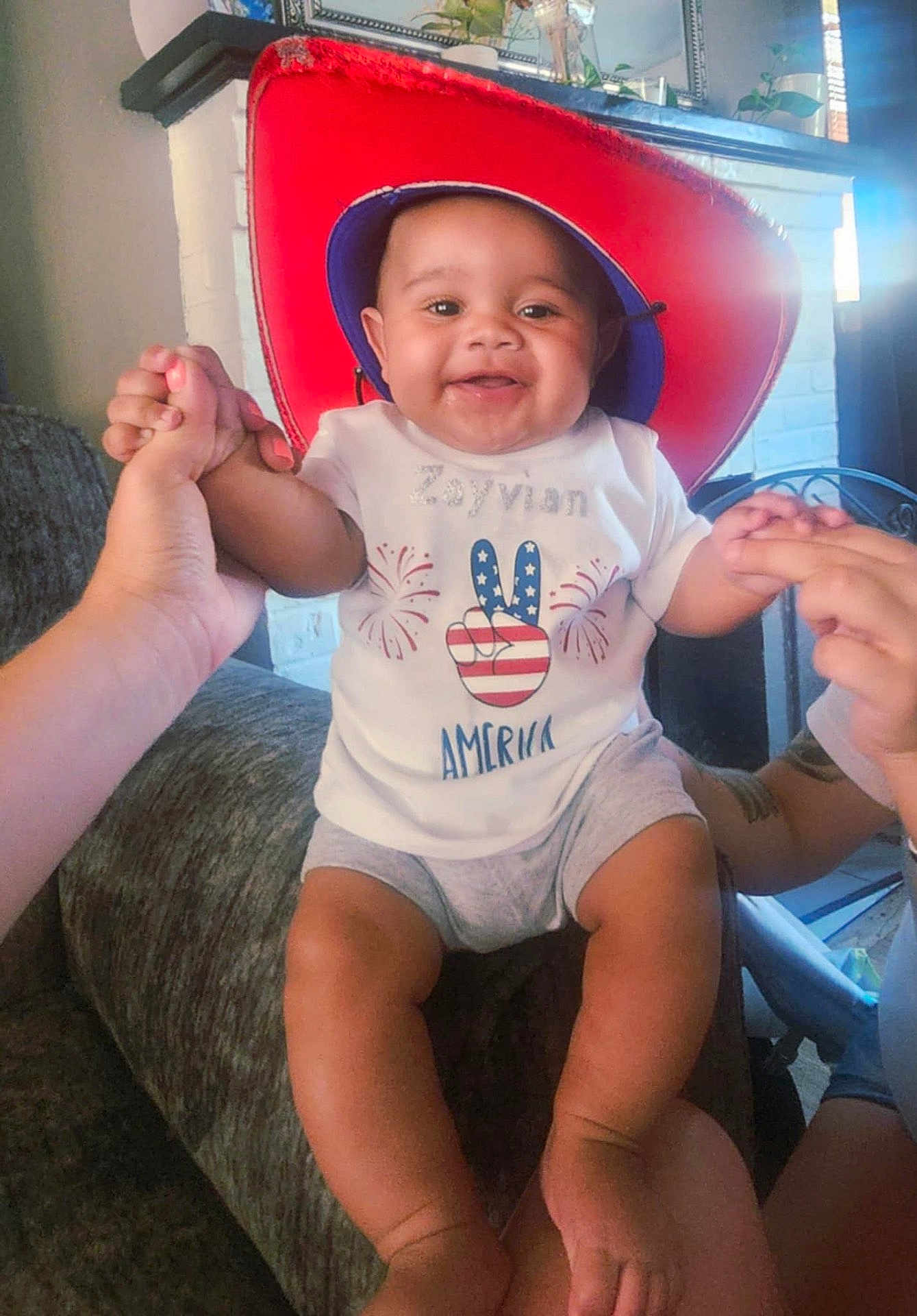 Zayvian joined the competition — help win amazing prizes! baby, child, smiling, hat, red_hat, shirt, american_flag, indoor, couch, holding_hands, person, happy, infant, home, furniture, legs, feet, toddler, celebration, peace_sign