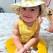 Crystal joined the competition — help win amazing prizes! child, toddler, smile, yellow_clothing, hat, floral_pattern, bed, bedspread, hand_holding, adult_hand, indoor, person, happy, face, sitting, cute, young_child, background, furniture, room