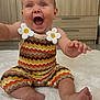 baby, child, smiling, happy, sitting, colorful_clothing, zigzag_pattern, flower_applique, barefoot, indoor, rug, wooden_cabinet, cute, excited, portrait, infant, playful, bright, cheerful, person