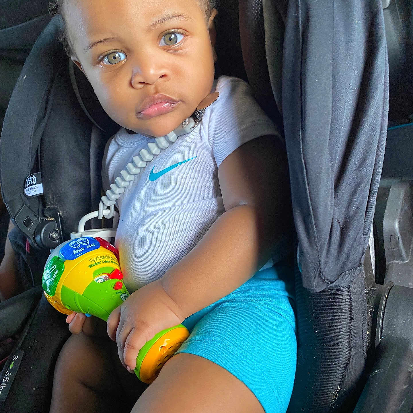 Keni joined the competition — help win amazing prizes! baby, blue_shorts, car_seat, child, colorful_toy, curious, cute, expression, green_eyes, hand, indoors, legs, nike_logo, person, portrait, safety, seatbelt, toddler, toy, white_shirt