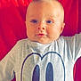 baby, blue_eyes, cartoon_shirt, child, clothing, cute, expression, face, indoors, infant, laying_down, light, looking_at_camera, person, portrait, red_background, shirt, smiling, toddler, young_child