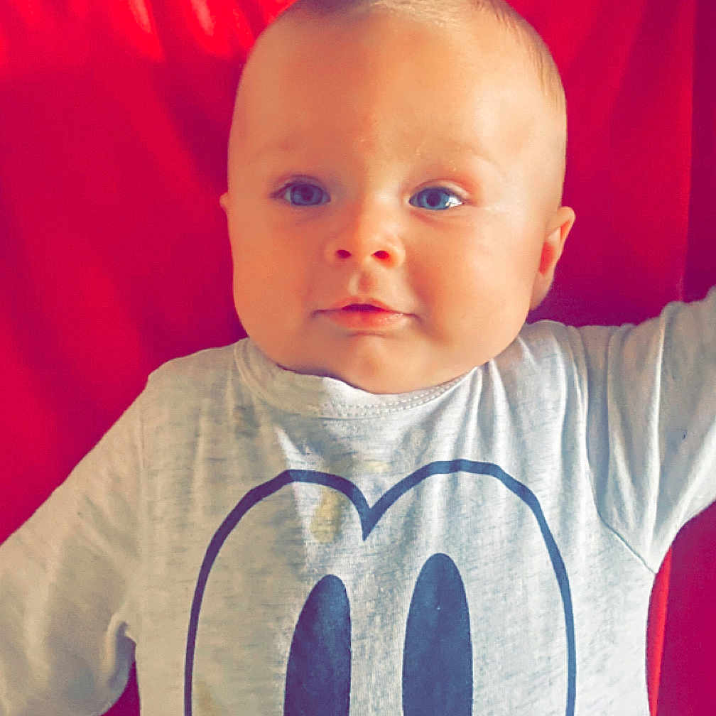 Kalvyn a rejoint le concours — aidez-le/la à gagner de superbes lots ! baby, baby_clothes, blue_eyes, child, closeup, cute, expression, face, head, indoors, infant, lying_down, mickey_mouse, person, playful, portrait, red_background, shirt, smiling, toddler