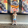 child, smiling, standing, movie_theater, posters, lilo_and_stitch, wall, carpet, tshirt, shorts, shoes, girl, indoor, happy, posing, hair, light, background, entertainment, display