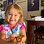 child, smiling, butterfly, monarch_butterfly, hands, colorful_clothing, indoor, table, chair, sports_memorabilia, framed_jersey, door, face, happy, person, young_child, home, wooden_furniture, natural_light, floor