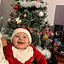 baby, child, christmas_tree, santa_claus_costume, holiday, smiling, tattoo, person, indoor, decorations, ornaments, tree_lights, festive, red_clothing, white_trim, happy, holiday_spirit, celebration, seasonal, holding