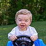 blond_hair, child, cute, daytime, fun, grass, greenery, happy, nature, outdoor, playtime, portrait, sitting, smiling, steering_wheel, sunlight, toddler, toy_car, white_shirt, young_child