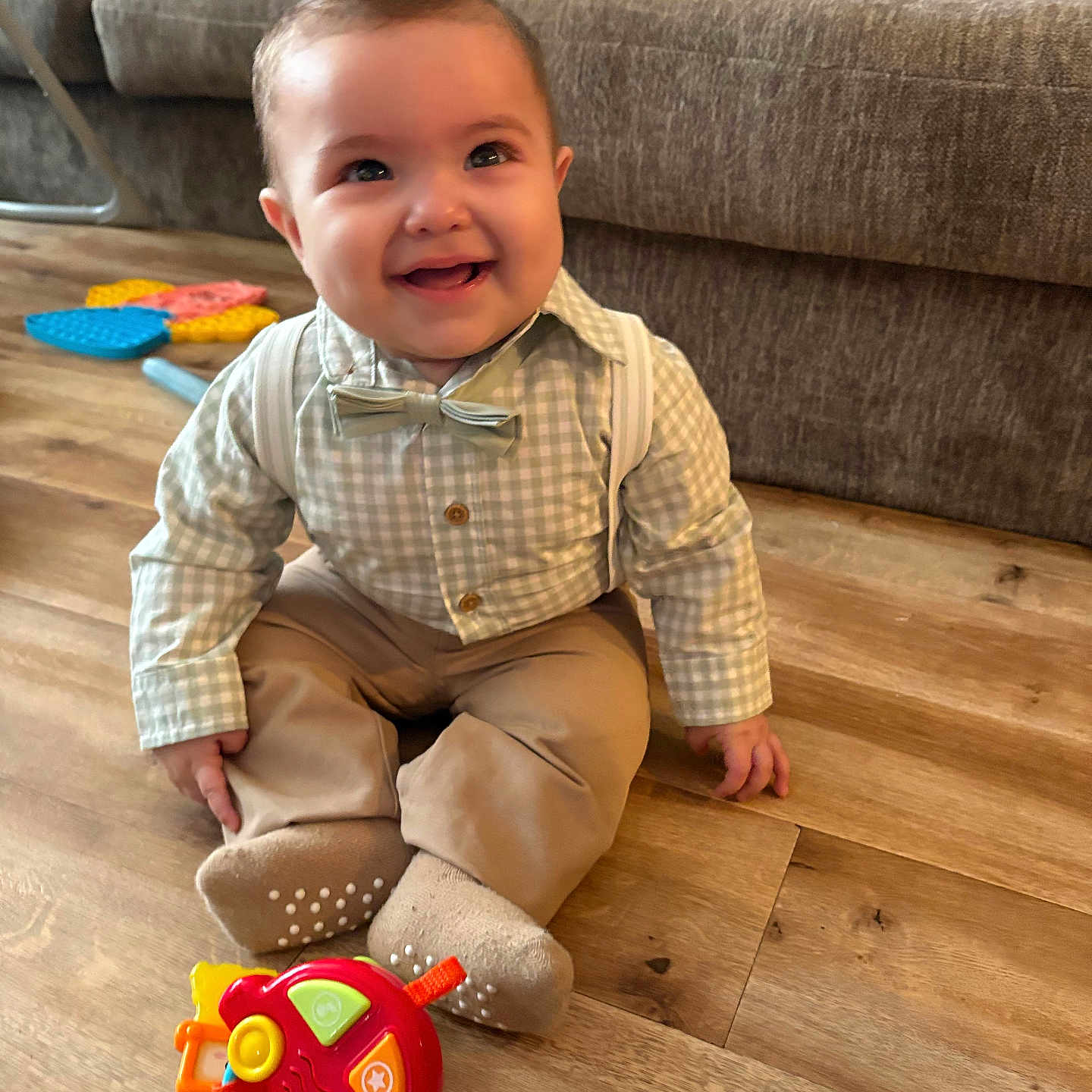 Ricardo joined the competition — help win amazing prizes! baby, child, smiling, indoor, floor, toy, car_toy, clothing, bow_tie, suspenders, wooden_floor, sofa, happy, cute, sitting, face, person, playful, living_room, infant