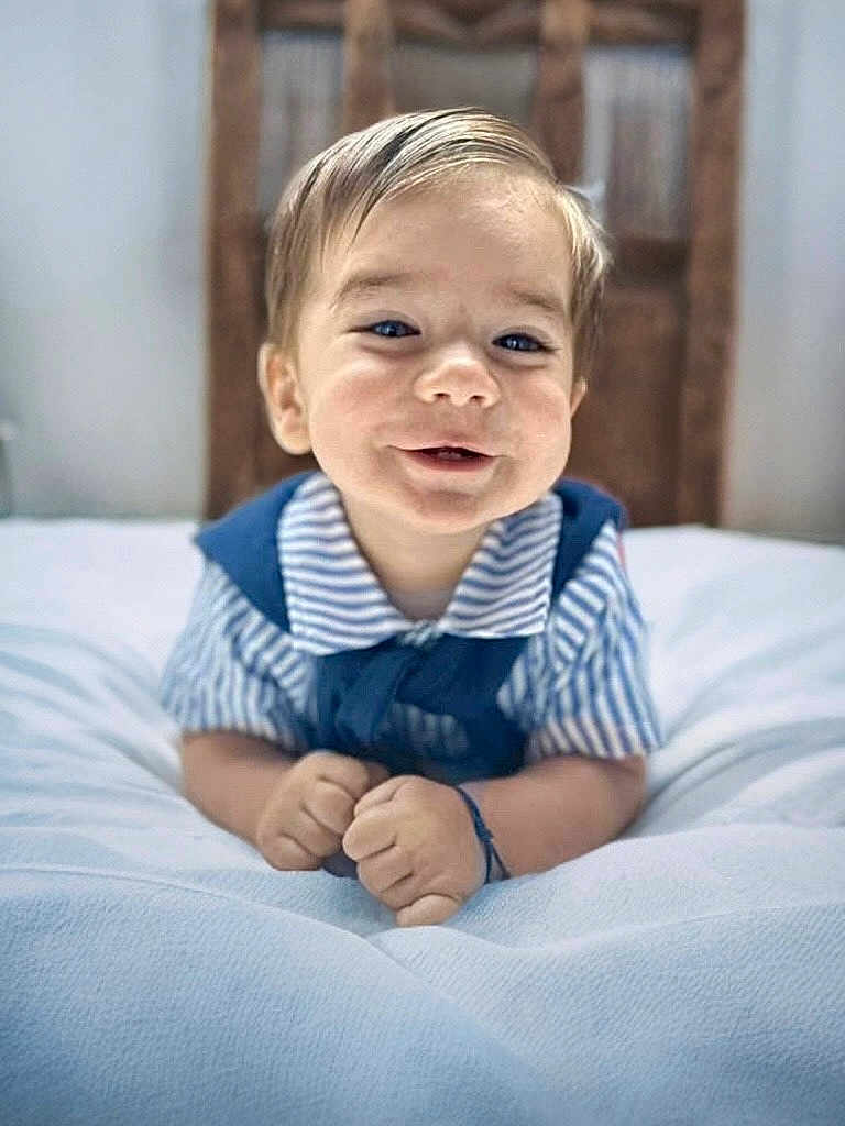 Mylan a rejoint le concours — aidez-le/la à gagner de superbes lots ! baby, child, smiling, portrait, indoor, bed, white_bedspread, striped_clothing, blue_clothing, hands, face, head, hair, happy, cute, infant, young_child, wooden_headboard, soft_texture, lying_down