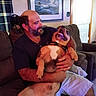Bandit is registered to the contest to win money with this photo: arm, beard, casual_clothing, couch, curtain, dog, door, furniture, hand, happy, home, indoor, living_room, man, pet, picture_frame, shorts, sitting, smiling, window