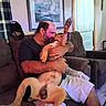affection, balding, beard, casual_clothing, couch, curtains, dog, home, indoor, living_room, love, man, painting, paw, person, pet, seated, shorts, smile, window