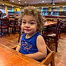 toddler, child, curly_hair, blue_tank_top, shark_design, high_chair, wooden_table, restaurant, dining_room, wooden_chairs, smile, indoor, ceiling_fans, lights, person, cute, furniture, happy, portrait, young_child