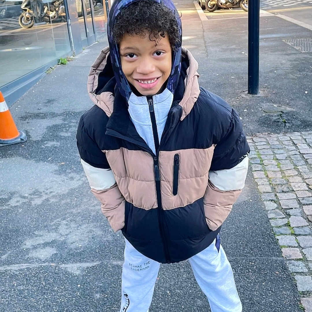 Iliandro participe au concours pour gagner de l'argent avec cette photo : child, boy, smiling, jacket, hoodie, sweatpants, sneakers, sidewalk, urban, street, outdoor, daylight, casual, happy, curly_hair, person, clothing, fashion, posing, city
