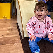 Khadija is registered to the contest to win money with this photo: toddler, child, smiling, pink_sweatshirt, cherry_pattern, blue_pants, socks, indoor, wooden_slide, rug, happy, playroom, flooring, hand, person, casual_clothing, play, fun, young_child, expression