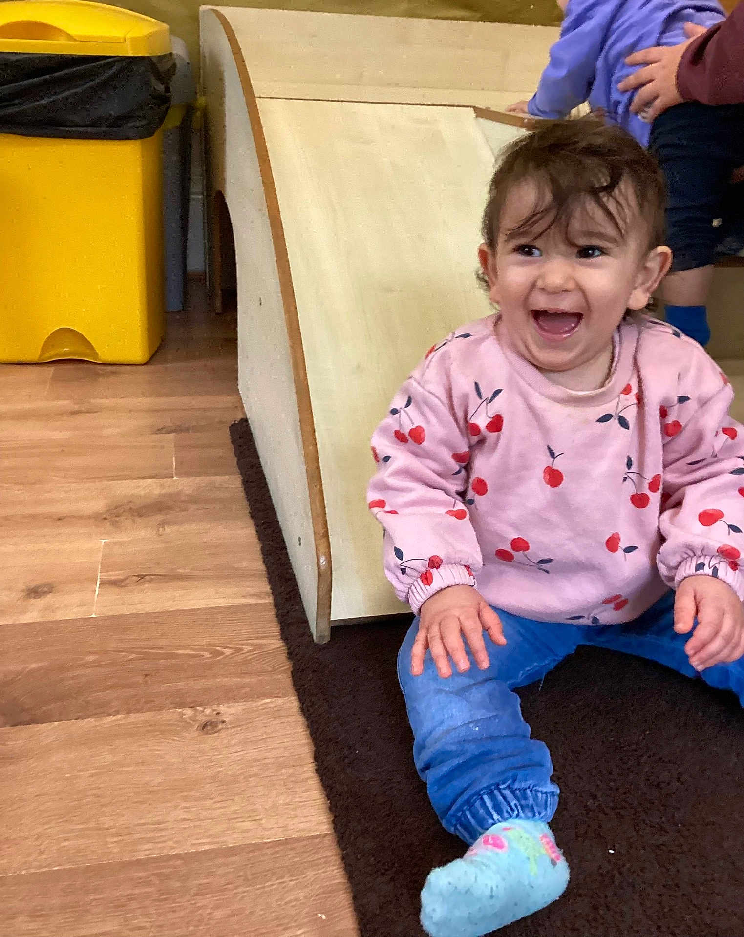 Khadija is registered to the contest to win money with this photo: toddler, child, smiling, pink_sweatshirt, cherry_pattern, blue_pants, socks, indoor, wooden_slide, rug, happy, playroom, flooring, hand, person, casual_clothing, play, fun, young_child, expression