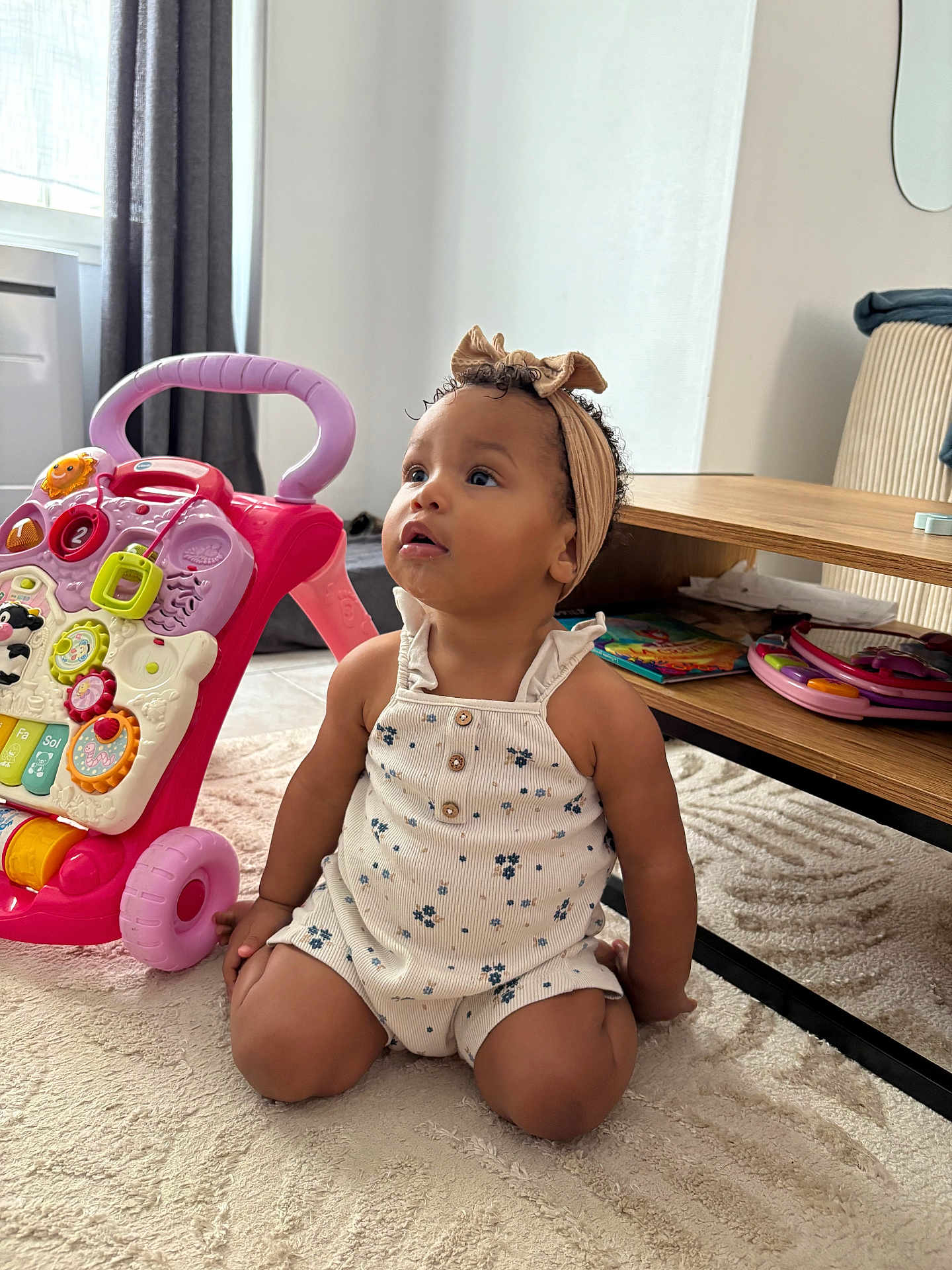 Alina participe au concours pour gagner de l'argent avec cette photo : toddler, child, baby, headband, romper, toy_walker, carpet, table, book, indoor, curtains, window, floor, furniture, person, sitting, playing, cute, curious, expression