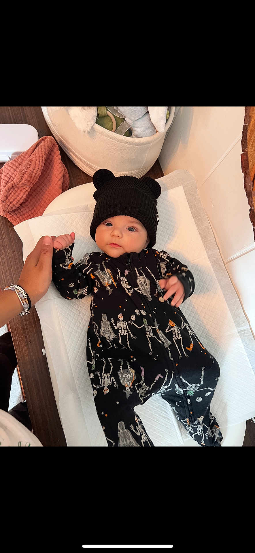 Kai joined the competition — help win amazing prizes! baby, infant, child, onesie, skeleton_pattern, black_hat, bear_ears, hand_holding, changing_pad, soft_toys, basket, pink_cloth, wooden_surface, indoor, cute, portrait, person, clothing, newborn, cozy