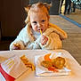 child, toddler, eating, fries, chicken_nuggets, fast_food, mcdonalds_logo, restaurant_interior, table, jacket, hooded_coat, pigtails, blonde_hair, blue_eyes, face, hands, portrait, meal, indoor, casual