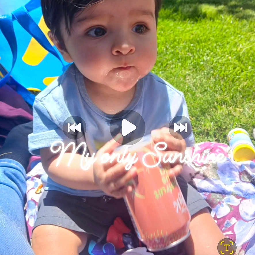 Benjamin Blue is registered to the contest to win money with this photo: baby, child, toddler, sitting, outdoors, sunlight, grass, picnic, blanket, toy, bottle, blue_chair, shorts, shirt, portrait, face, wide_eyes, dark_hair, playtime, text_overlay