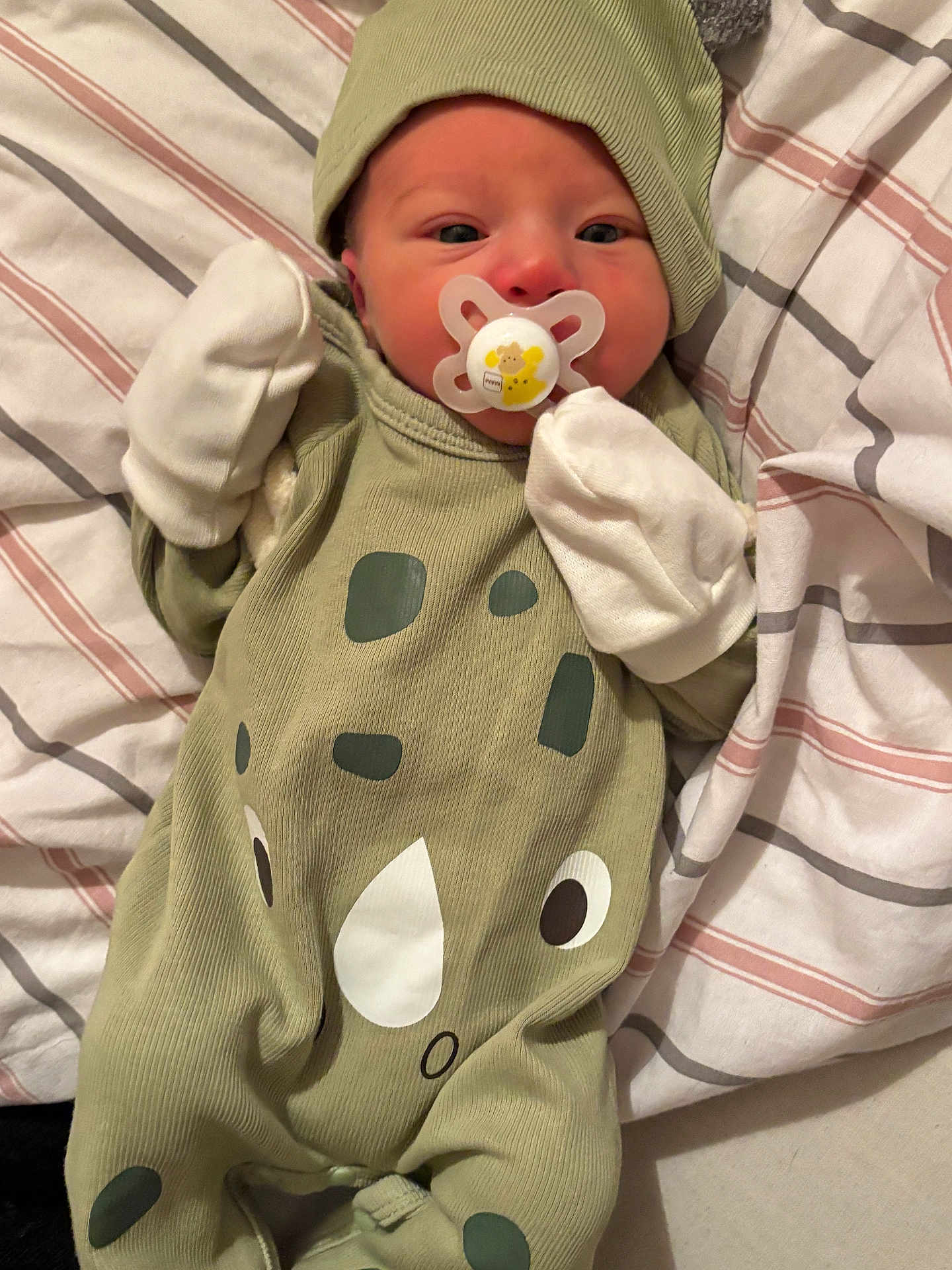 Saturn is registered to the contest to win money with this photo: baby, newborn, pacifier, mittens, hat, green_outfit, rhino_design, striped_blanket, lying_down, infant, sleepwear, cute, small, child, person, cozy, soft, indoor, resting, wearing