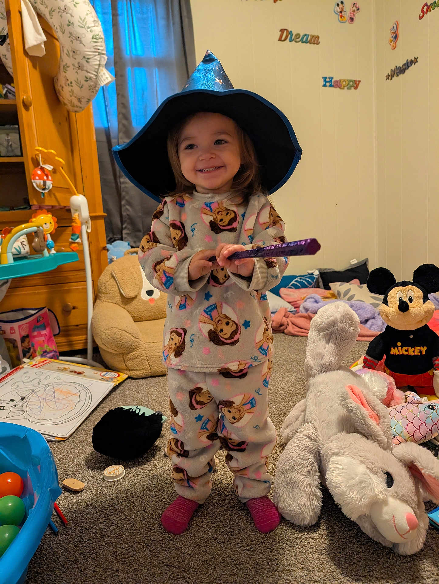 Brynlee is registered to the contest to win money with this photo: toddler, child, pajamas, wizard_hat, wand, smiling, toy_room, stuffed_animals, plush_bunny, mickey_mouse, carpet, coloring_book, toy_balls, furniture, curtains, wall_decor, happy, indoor, playful, person
