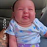 baby, child, tongue_out, smiling, floral_clothing, purple_shorts, white_shirt, car_seat, tattooed_arm, hand, infant, cute, portrait, indoors, person, seat, holding, playful, expression, date_stamp