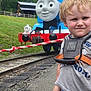 Colton is registered to the contest to win money with this photo: child, toddler, train, thomas_the_tank_engine, railroad_tracks, outdoor, grass, sky, cloudy, jeans, tshirt, safety_harness, rope, fence, smile, blond_hair, person, playful, portrait, daytime