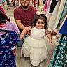 child, toddler, adult, father, daughter, smiling, dress, white_dress, bow, fashion, clothing_store, clothing_racks, shopping, portrait, family, indoor, standing, sitting, accessories, happy