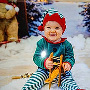 Eden a rejoint le concours — aidez-le/la à gagner de superbes lots ! baby, child, toy, wooden_toy, airplane_toy, elf_costume, striped_pants, red_hat, smiling, indoor, holiday_theme, winter_background, teddy_bear, snow, festive, cute, playful, sitting, portrait, happy