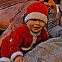 Tiago a rejoint le concours — aidez-le/la à gagner de superbes lots ! baby, child, santa_suit, red_clothing, hat, smiling, indoor, couch, adult_hand, holiday, festive, cozy, warm_lighting, person, face, clothing, happy, infant, celebration, family