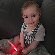 Bobby is registered to the contest to win money with this photo: baby, child, infant, sitting, couch, blanket, striped_overalls, white_shirt, glowing_toy, red_light, smiling, curious_eyes, indoors, soft_lighting, cute, happy, person, face, legs, hands