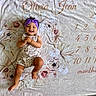 baby, child, smiling, purple_headband, romper, floral_pattern, blanket, name_text, numbers, months, happy, infant, cute, clapping_hands, lying_down, indoor, portrait, skin, face, head