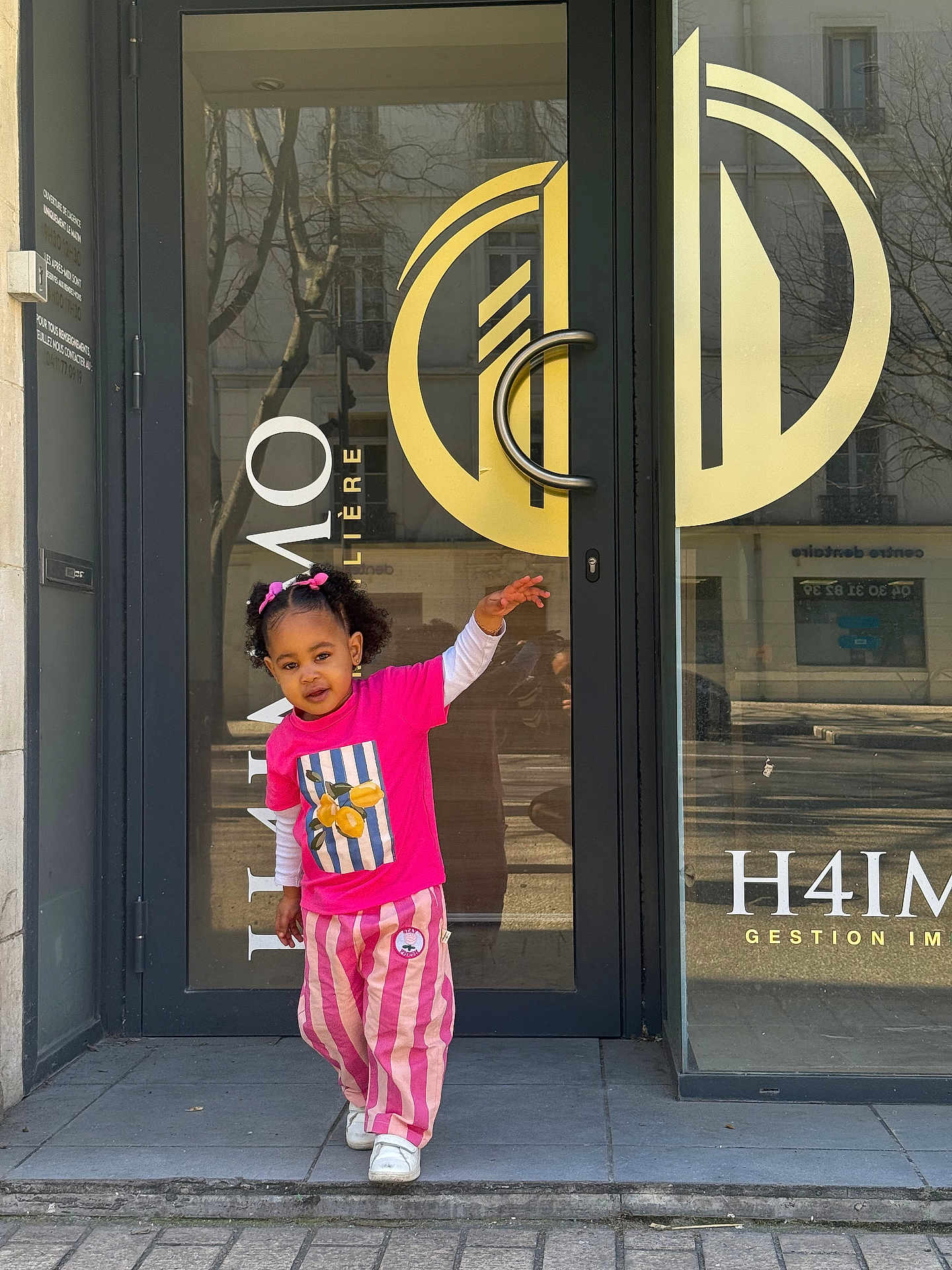 Elyjah a rejoint le concours — aidez-le/la à gagner de superbes lots ! child, toddler, girl, pink_clothing, striped_pants, glass_door, storefront, logo, door_handle, reflection, sidewalk, urban, portrait, standing, white_sneakers, hair_bows, smile, window, shadow, outdoor