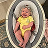 baby, smiling, yellow_onesie, headband, baby_rocker, striped_cushion, infant, happy, child, indoor, floor, carpet, footwear_absent, cute, playful, person, home, comfort, seated, portrait