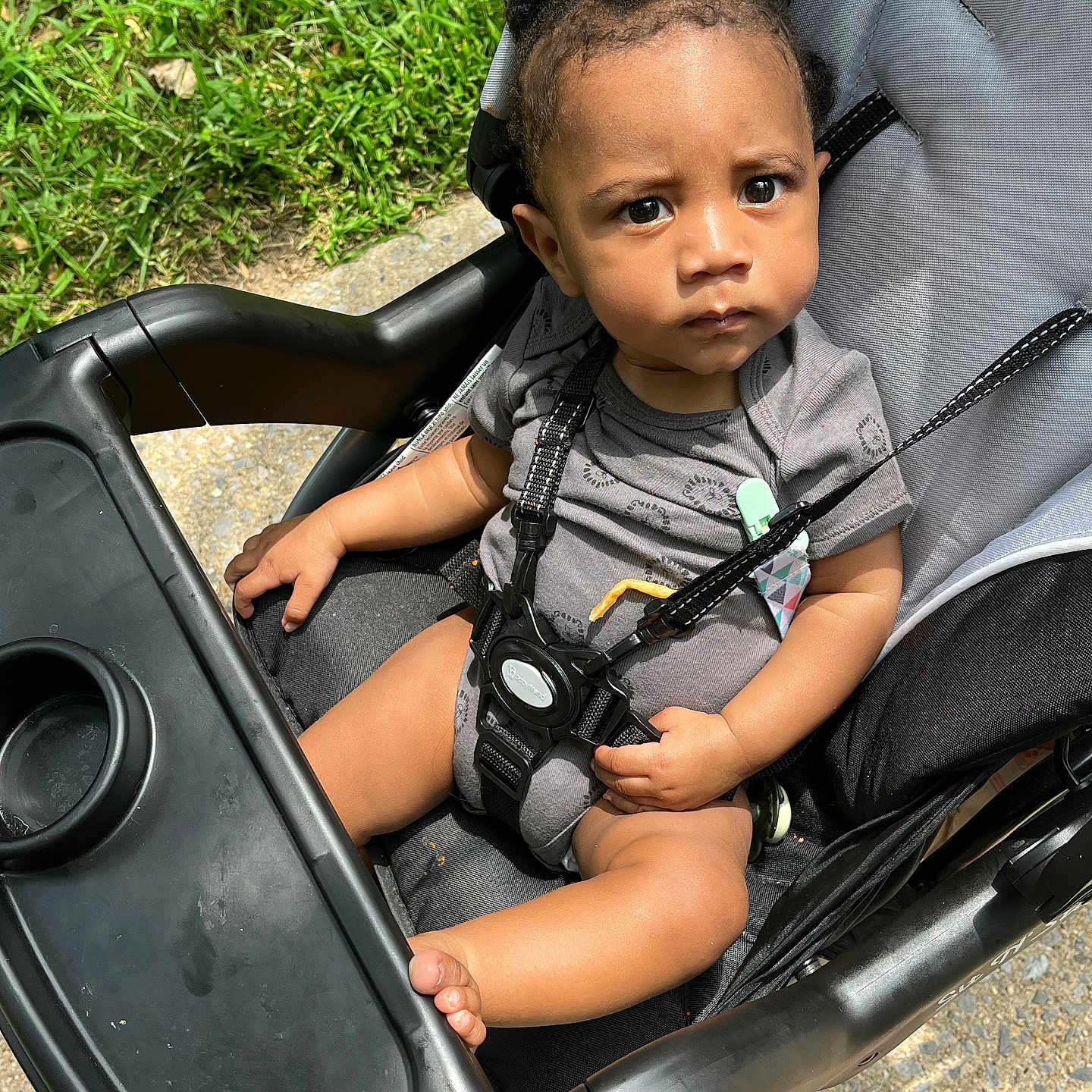 Today joined the competition — help win amazing prizes! baby, baby_boy, black_plastic, child, closeup, curly_hair, daylight, foot, grass, hand, harness, infant, onesie, outdoor, pavement, person, seat, serious_expression, stroller, sunlight