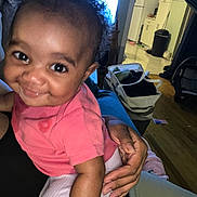 Tomorrow is registered to the contest to win money with this photo: baby, child, smiling, curly_hair, indoor, person, hand, clothing, pink_shirt, white_pants, living_room, kitchen, furniture, basket, floor, home, holding, cute, happy, face